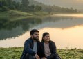 Couple sitting by Mulshi Lake at sunrise with hills reflecting in calm water, warm golden light, wide peaceful landscape