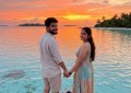 Indian couple standing on wooden jetty over turquoise Maldives lagoon at sunset, warm golden light, wide angle, natural