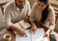 Couple comparing hotel bill and homestay receipt at wooden table, natural morning light, warm tones, overhead flat lay s