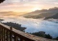 Panoramic view of Mulshi Lake at sunrise with mist rolling over calm blue water, shot from a homestay balcony with woode