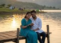Couple sitting by Pawna Lake at sunset, golden light reflecting on calm water, wide scenic view, warm and peaceful mood