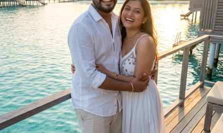 Indian couple standing on overwater villa deck, turquoise lagoon below, golden hour light, candid smiles, natural unpose